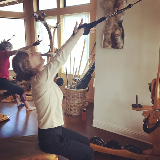 Woman using Pilates equipment, arms raised, in a light-filled studio.