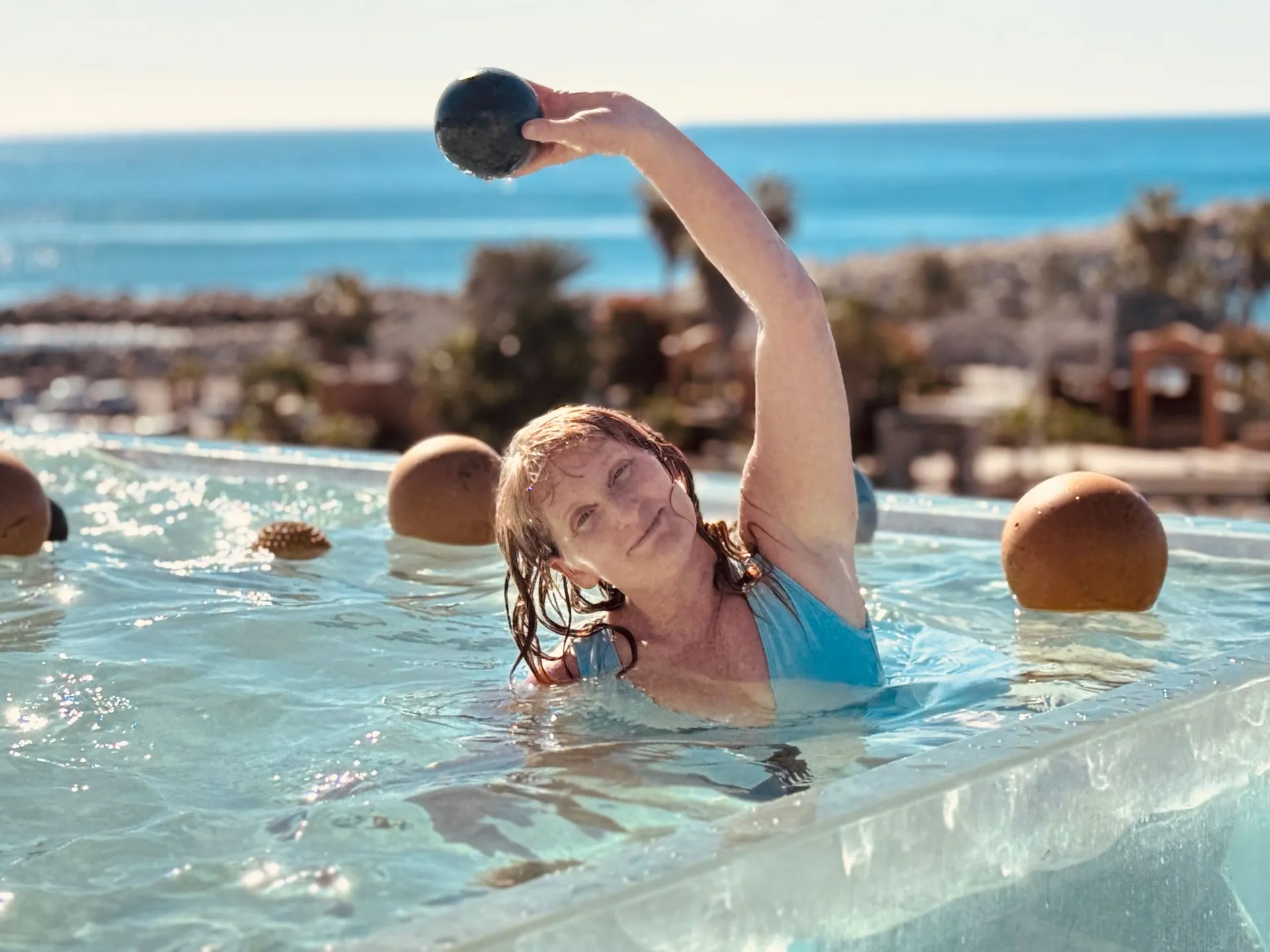 Woman in a pool raises a ball overhead; ocean and landscape in the background.