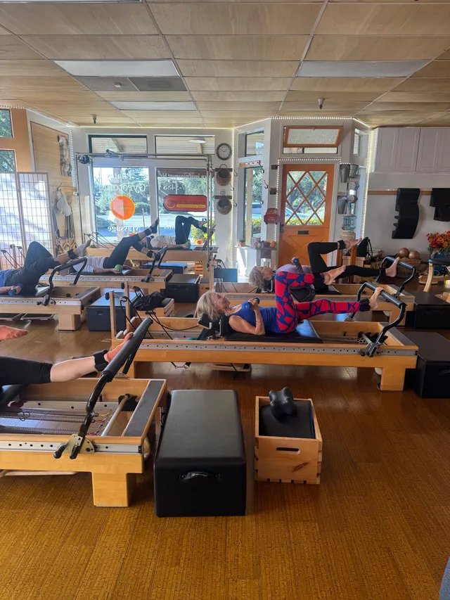 Pilates class in a studio; people on reformer machines, exercising. Sunlight streams through the windows.